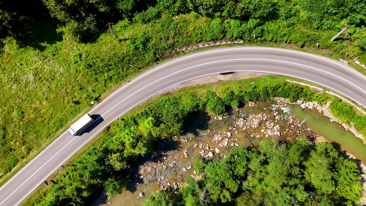 Truck on a winding road by stream. A large truck travels along a winding road surrounded by lush greenery and a stream on a sunny day