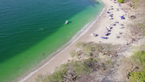 Popular-Sandy-Beach-Shore-With-Tents-And-Boat-On-Ocean,-Playa-Conchal-Costa-Rica,-4K-Drone