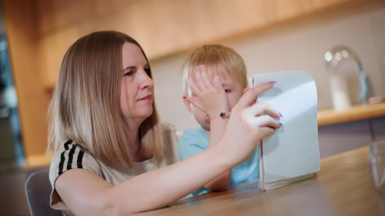 Woman sitting with child at wooden table holding open notebook while boy grips pen with serious face, learning activity, guidance, support, encouragement, concentration, development, home environment
