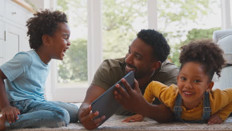 father at home with children lying on rug playing with digital tablet
