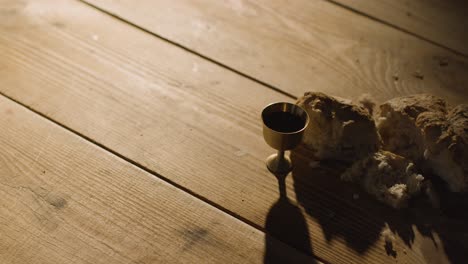 concepto religioso filmado con cáliz pan roto y vino en una mesa de madera con un charco de luz 1