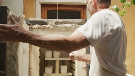 Senior-caucasian-man-wearing-apron-and-firing-pottery-in-kiln-at-pottery-workshop