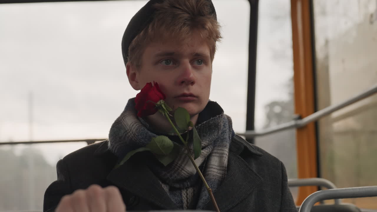 Melancholic white man on tram, rose. Young white poet creator clutching red rose, fogged window and rain, wool scarf and beanie, tight grip on metal rail, intimate cinematic portrait, moody winter