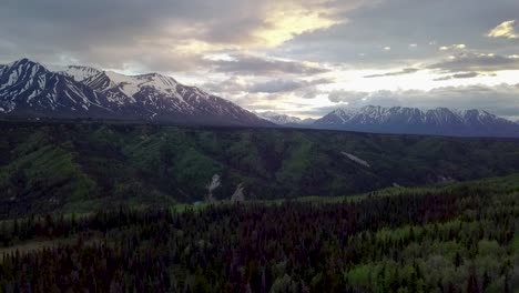 beautiful countryside scenic flight above yukon green lush tree forest with snow capped mountain range in background on dramatic cloudy day, canada, overhead aerial sideways descend