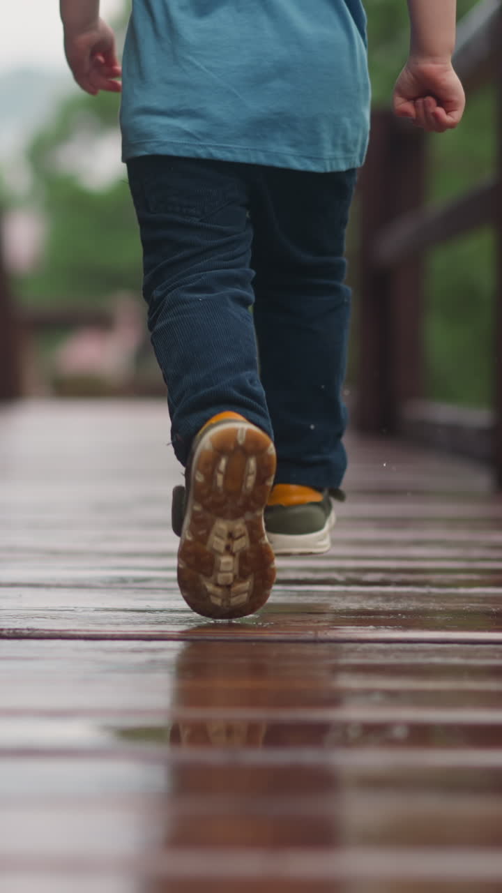 Playful toddler boy in sneakers runs along wet after rain veranda deck with railing at wooden cottage house on spring day close backside view