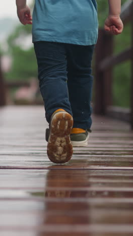 playful toddler boy in sneakers runs along wet after rain veranda deck with railing at wooden cottage house on spring day close backside view
