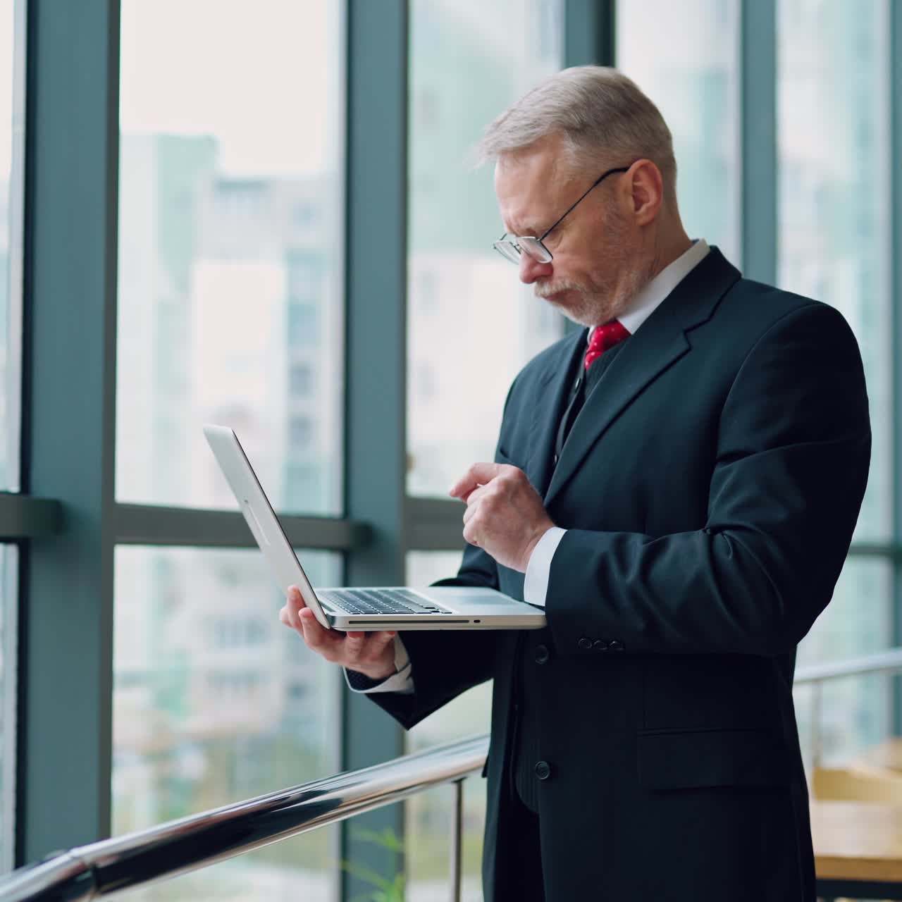 Mature senior serious businessman with laptop in hands thinking while checking e-mails standing near window with panoramic city view. Video from the side.