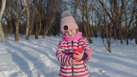 a young girl in a pink hat and scarf smiles as she looks at her phone in a snowy forest.