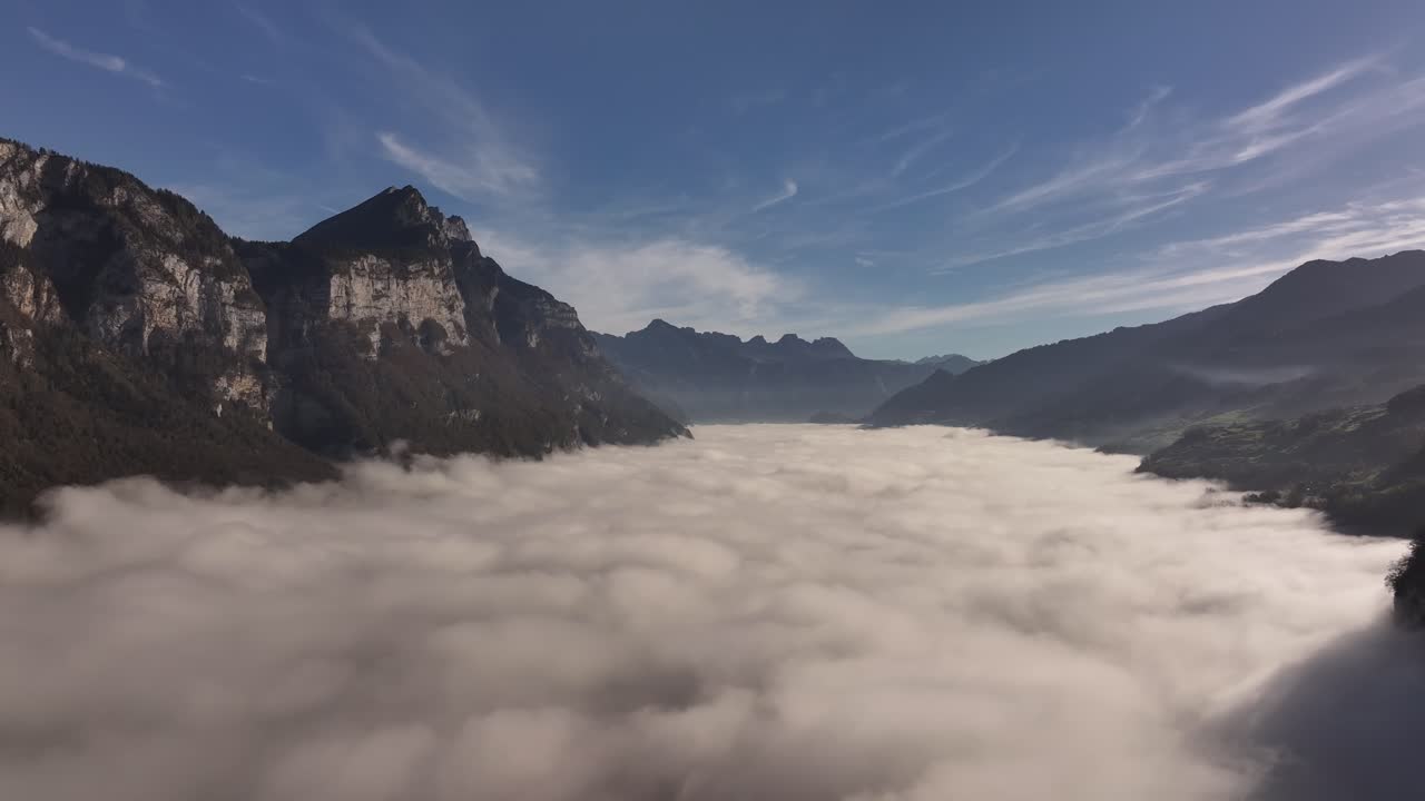 Clouds blanket a mountain valley under a bright blue sky with rugged peaks on either side