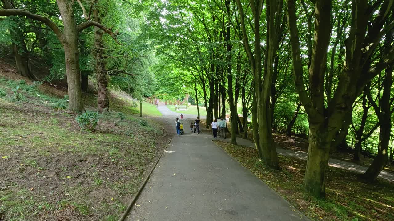 Several people walk along a paved path through lush, green botanical gardens. Daylight filters through dense trees, with steady camera movement and tranquil atmosphere