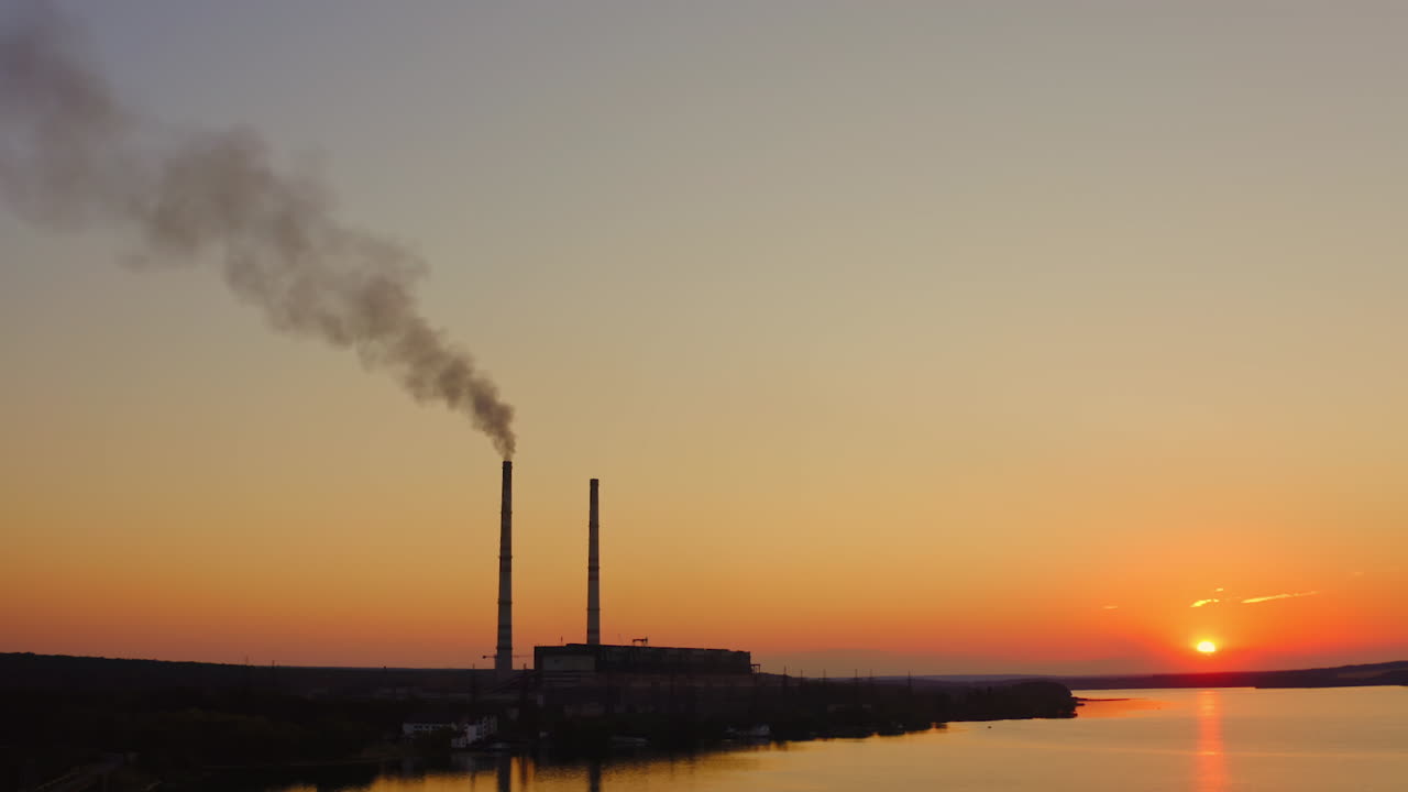 Plant with smoke against orange sky. Industry among nature at sunset. Harmful smoke released from factory in the air.