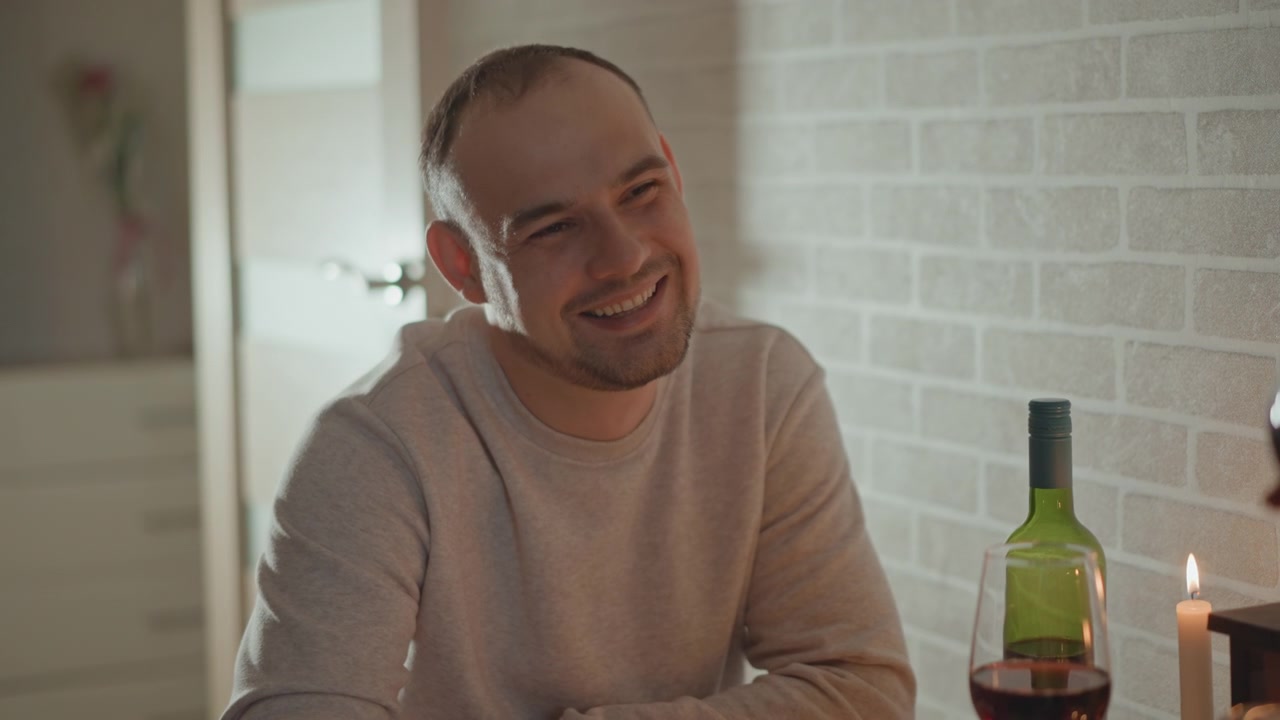 Giovane uomo seduto accanto al tavolo da pranzo al chiuso sorride e ride mentre reagisce a una conversazione non vista, con luce soffusa dalla finestra, mani rilassate e un'atmosfera sociale rilassata in un accogliente ambiente domestico moderno.