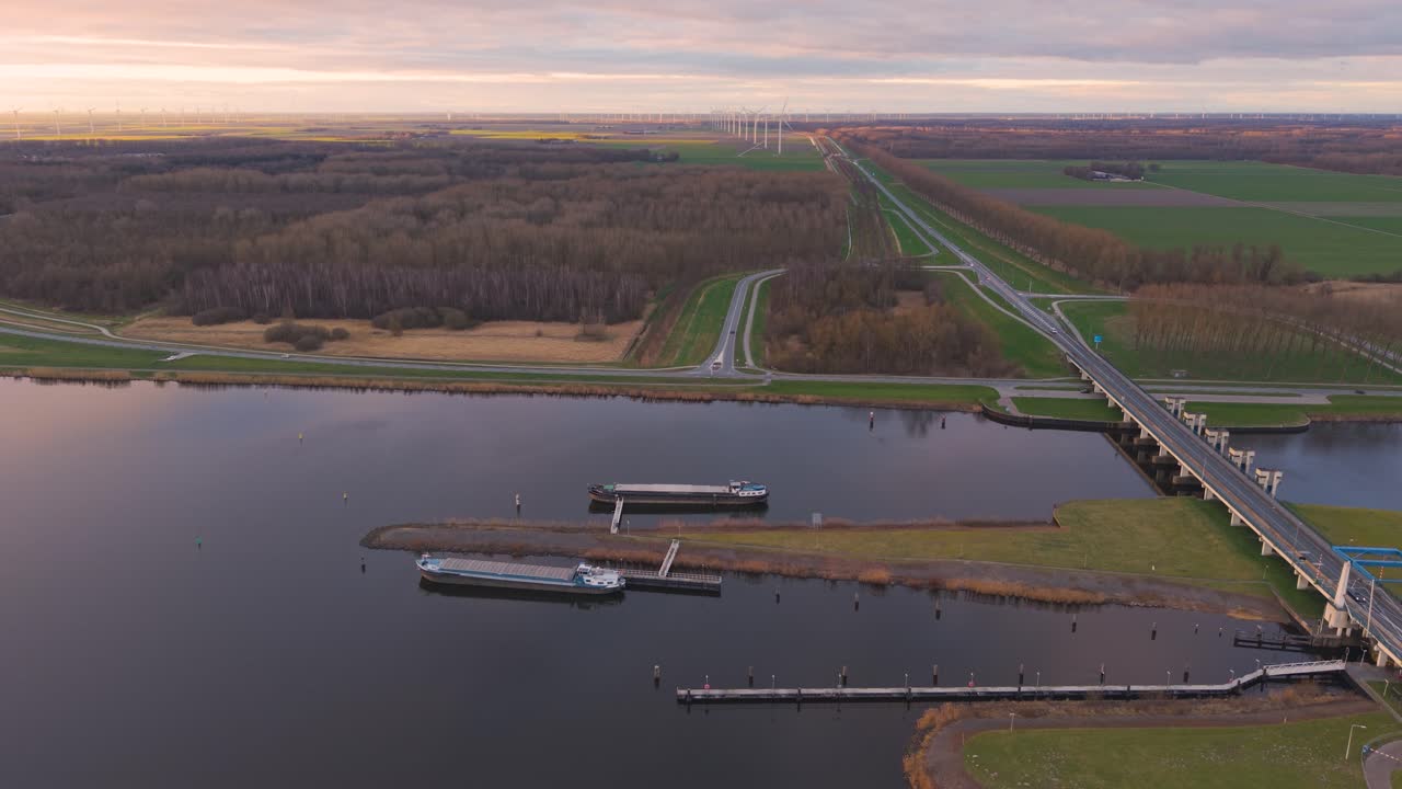 Drone view of a river, dam, bridge, and boats with roads, fields, and wind turbines in the background at sunset.