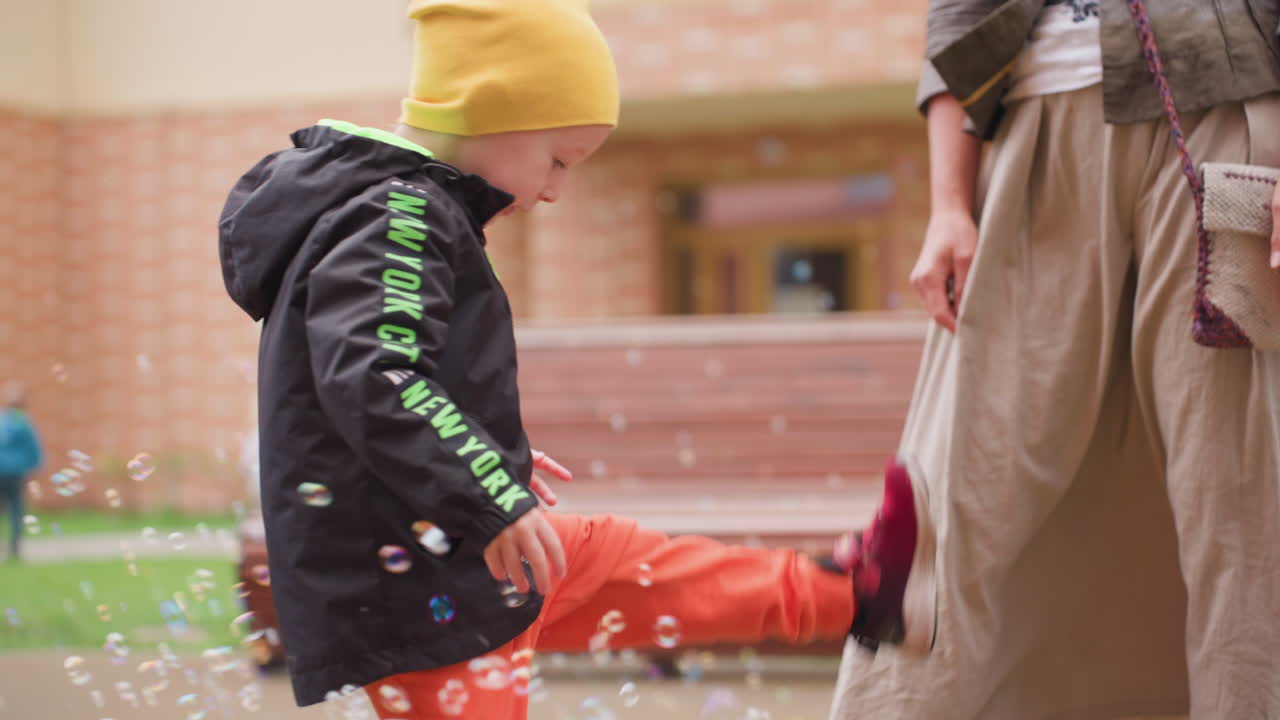 Young boy in yellow cap and black jacket playfully kicks floating soap bubbles while adult stands beside watching on sunny courtyard, joyful moment showing energy
