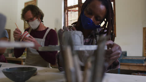 diverse male and female potters wearing face mask and apron working on pottery at pottery studio