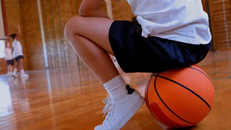 Low-section-Caucasian-schoolgirl-sitting-on-basketball-in-basketball-court-at-school-4k