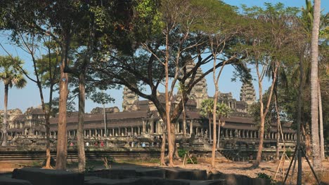 Wide-Exterior-Still-Shot-of-The-Corner-Of-Angkor-Wat-Temple-Through-Some-Trees-with-far-Away-Tourists-Outside-in-the-daytime