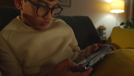 niño sentado en el sofá en casa jugando o transmitiendo a un dispositivo de juego portátil por la noche 2