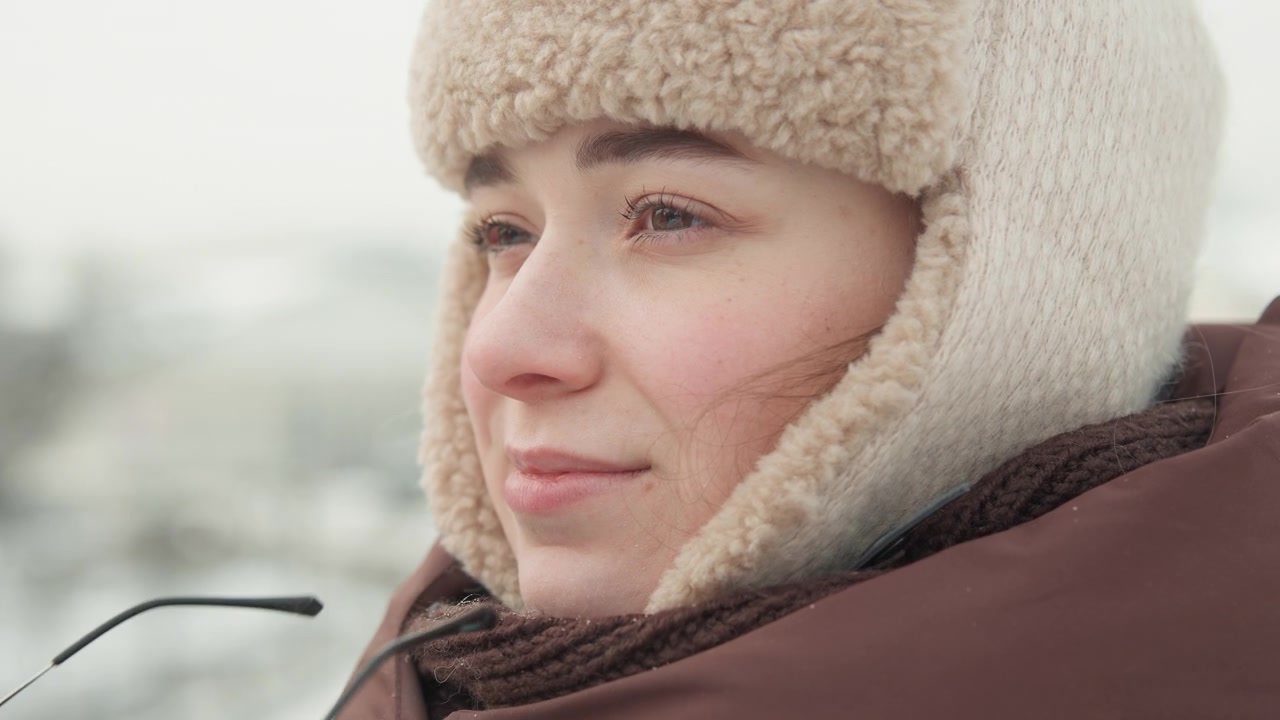 Close-up motion portrait of young person in shearling hat with earflaps and padded jacket, using both hands to adjust metal-rimmed glasses before turning toward snowy distance in soft daylight