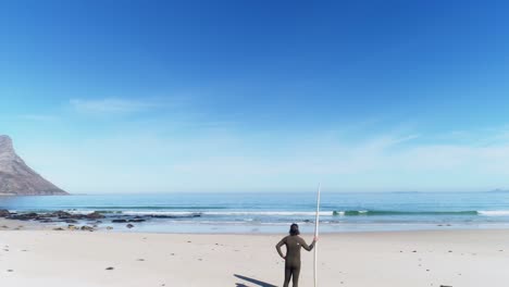 surfer standing alone on beach with surfboard next to him looking out over calm waters