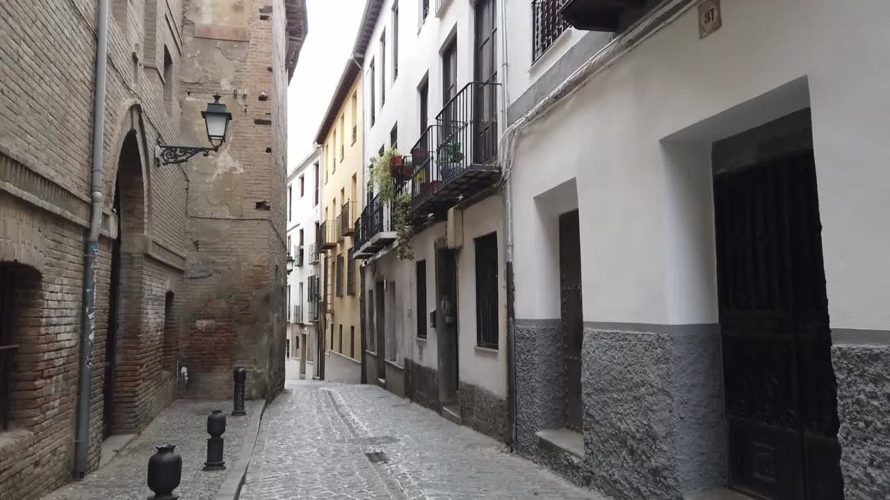 Young woman strolls narrow cobblestone alley in Granada's historic quarter. Colorful old buildings, lanterns, balconies. Authentic Spanish city exploration footage