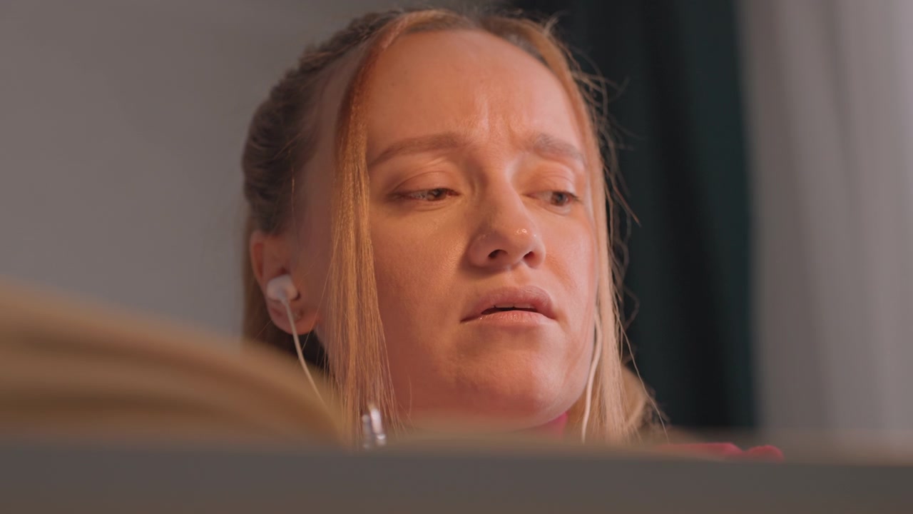 Close up of young woman with blonde hair tied back wearing. Her head moves slightly as woman focuses. Scene is indoors against plain gray wall with dark curtains