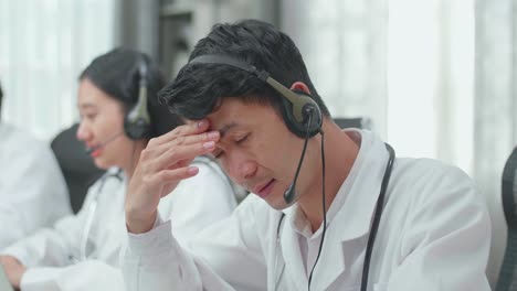 close up of an asian man doctor wearing headsets working as call center agent is tired due to working while his colleagues are speaking and typing during a call at the office