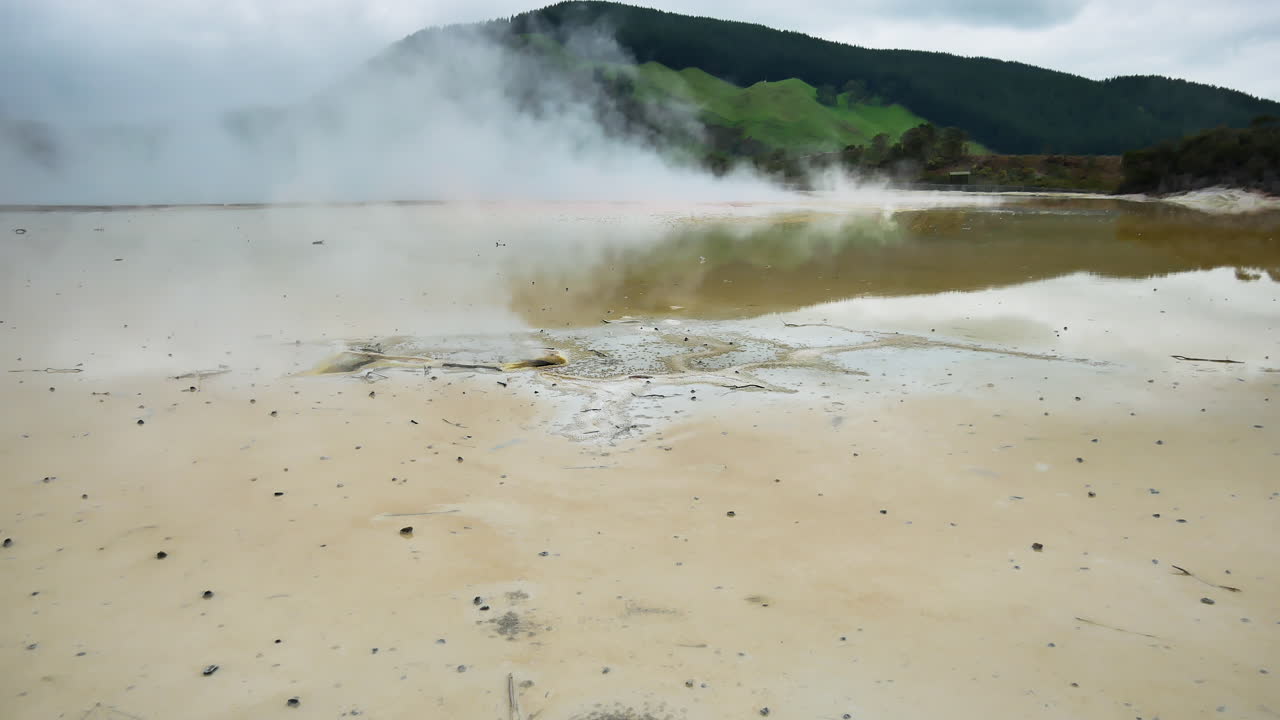 Panning shot of steam rising up from boiling water lakes in volcanic zone - Beautiful mountains in background during cloudy day - Wai-o-tapu in New zealand