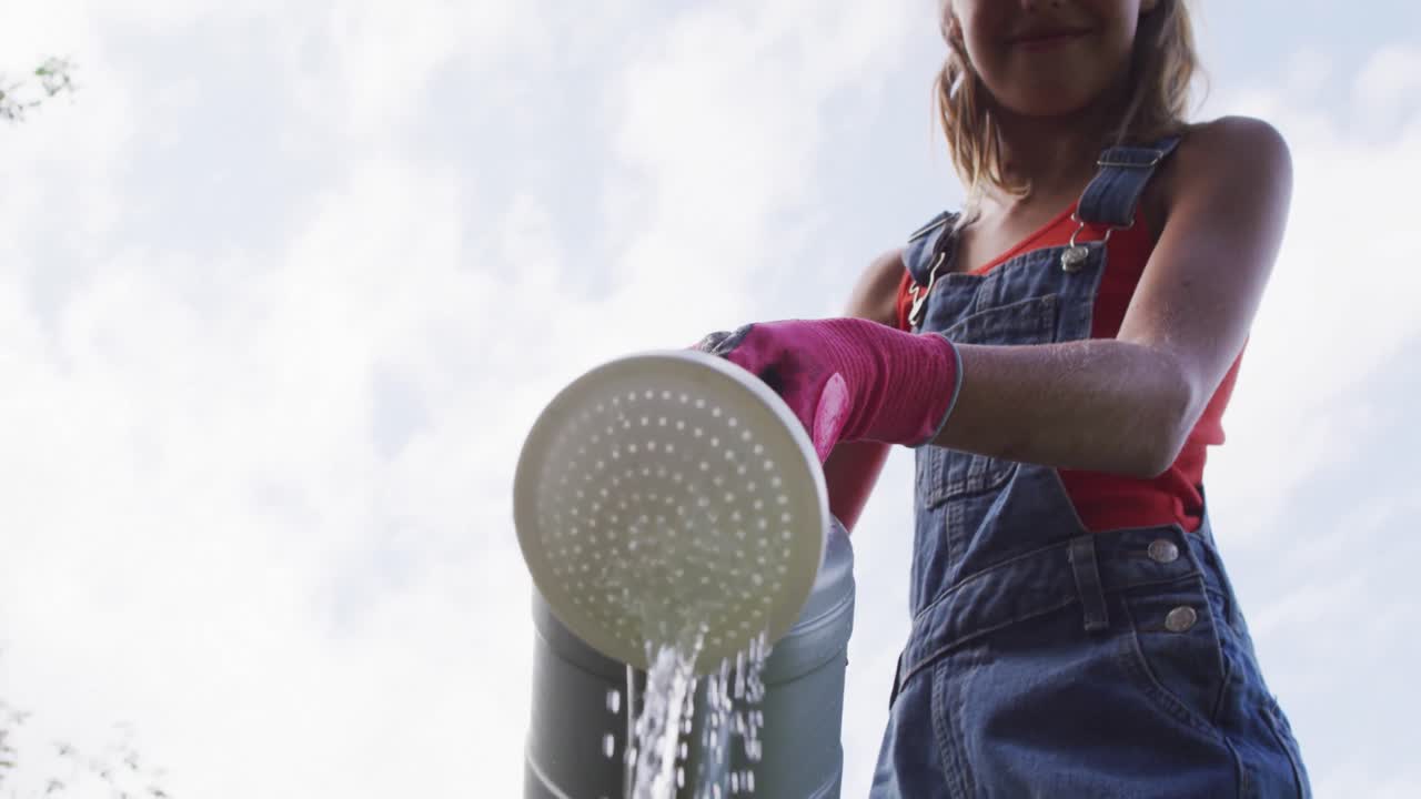 vista de bajo ángulo de una niña caucásica sonriente en el jardín regando plantas con un bote de riego