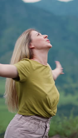 cheerful woman tourist with blonde hair spins around over large valley against distant hills at wild eco resort on gloomy day slow motion