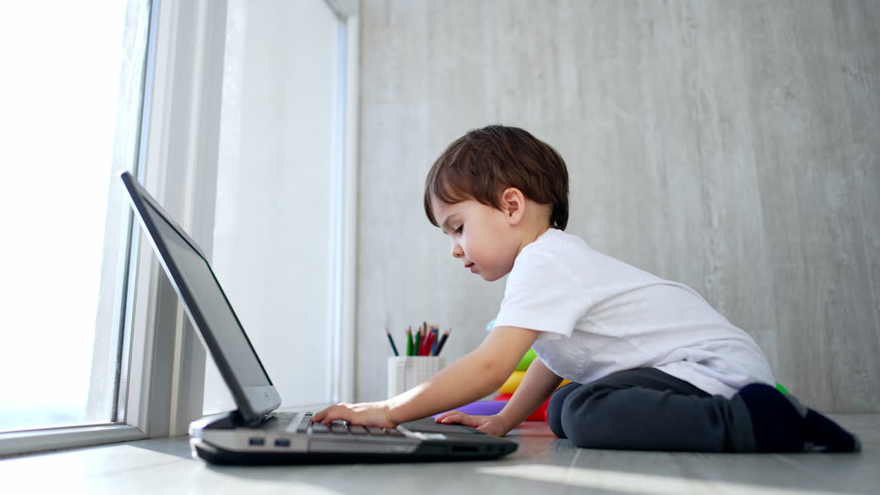 A child sits on the floor and looks at the laptop. A toy is on the table.