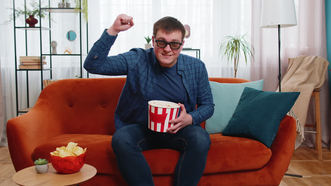 hombre emocionado con gafas 3d sentado en el sofá comiendo bocadillos de palomitas de maíz y viendo una película de televisión interesante
