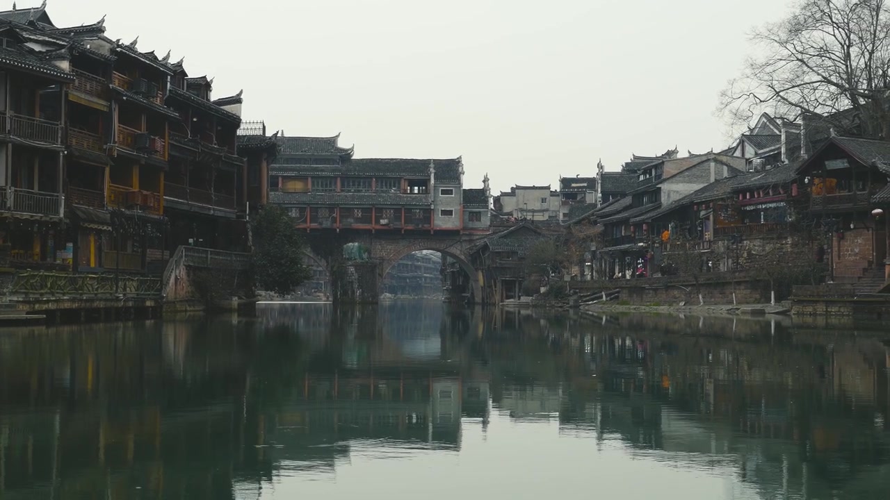 Um rio tranquilo flui através da Cidade Antiga de Fenghuang, refletindo casas tradicionais sobre palafitas e uma ponte coberta histórica, capturando o encanto deste tesouro cultural