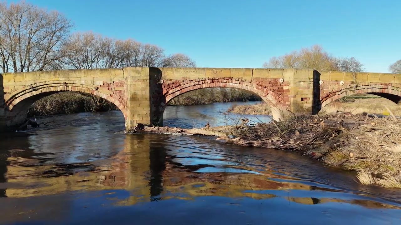 Ripresa aerea dal basso di uno storico ponte in pietra ad arco su un fiume che scorre, con riflessi sull'acqua e alberi spogli invernali sotto un cielo azzurro limpido
