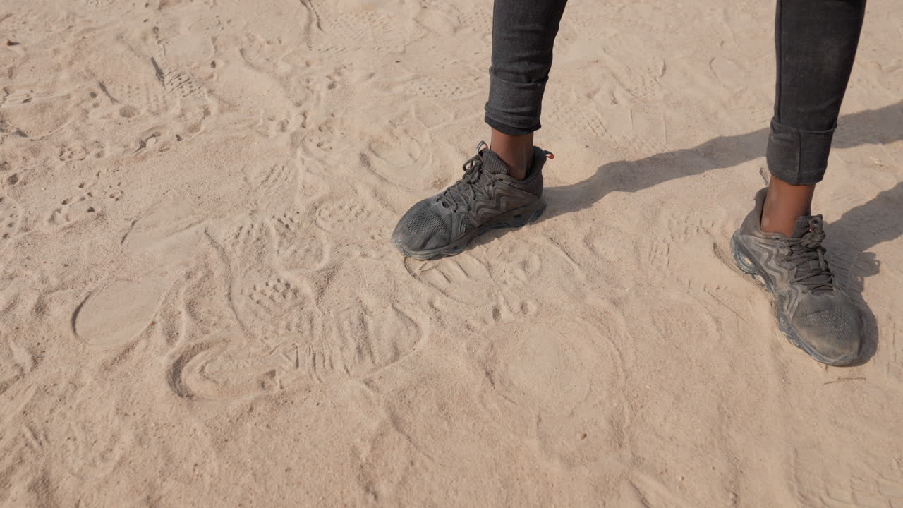 Kumlu zeminde siyah koşucunun ayakları, dokulu spor ayakkabılar ve tozlu taytlarla spor ısınma duruşu, gölgeli ayak bilekleri ve taze ayak izleri, parlak güneş ışığı altında sprint ve antrenman tatbikatlarına hazır.