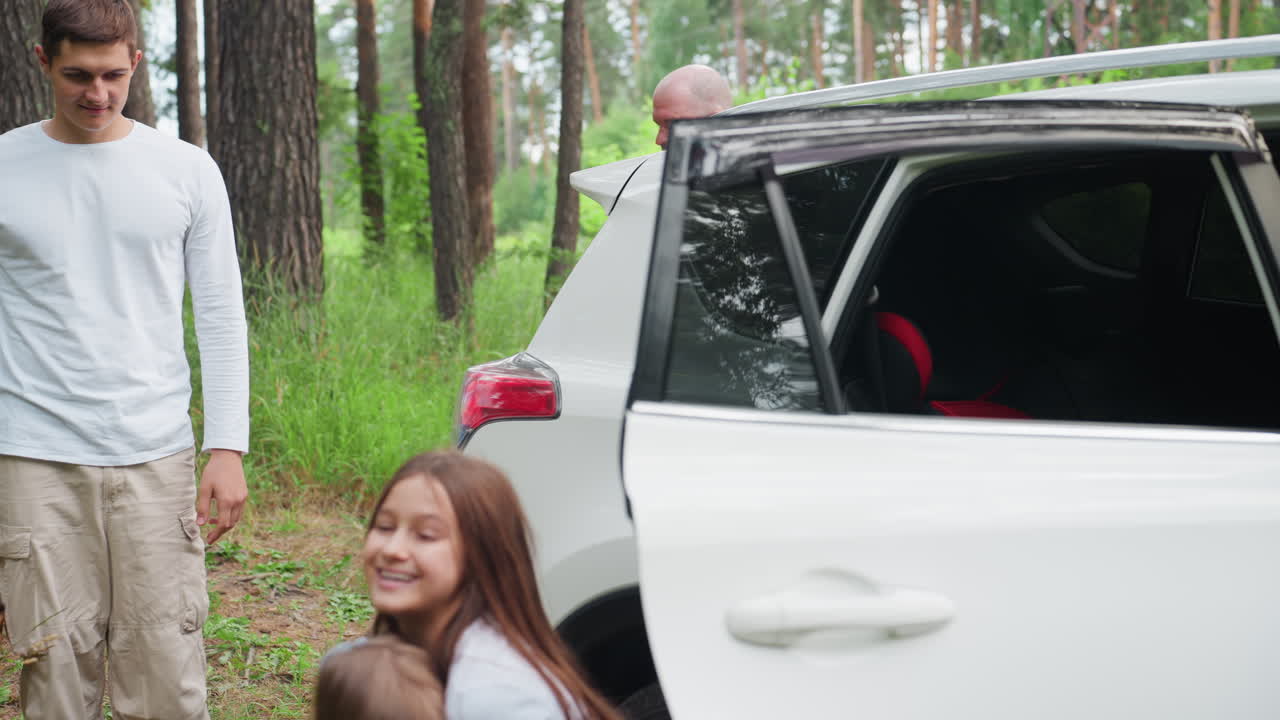 Una familia llega a un parque recreativo. Una niña ayuda a su hermana a bajar de un coche blanco, mientras su hermano y sus padres, que están cerca, se preparan para un picnic con rostros alegres, rodeados de un bosque verde.