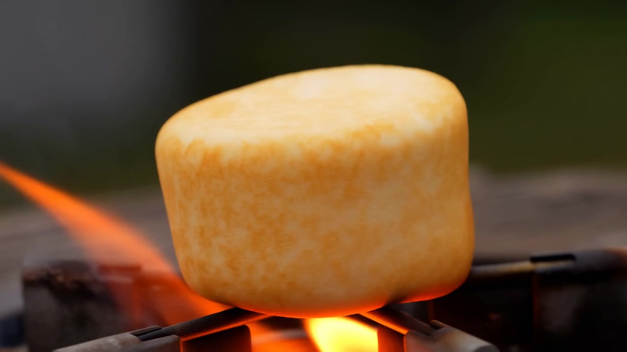 Nærbilde av et hvitt sylindrisk skumstykke grepet med metalltenger og varmet over en lys oransje flamme, med en mørk uskarp bakgrunn og varm verkstedatmosfære.
