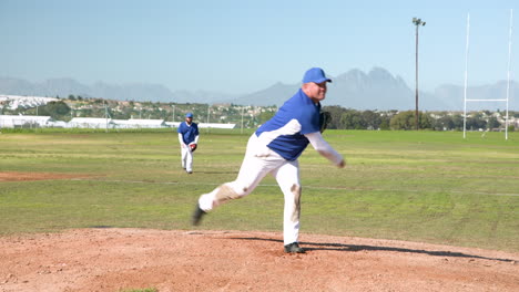 Pitching on baseball field, man in blue uniform holding glove, teammate watching