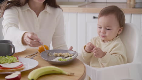 madre irreconocible alimentando a su linda niñita sentada en una silla alta en la cocina