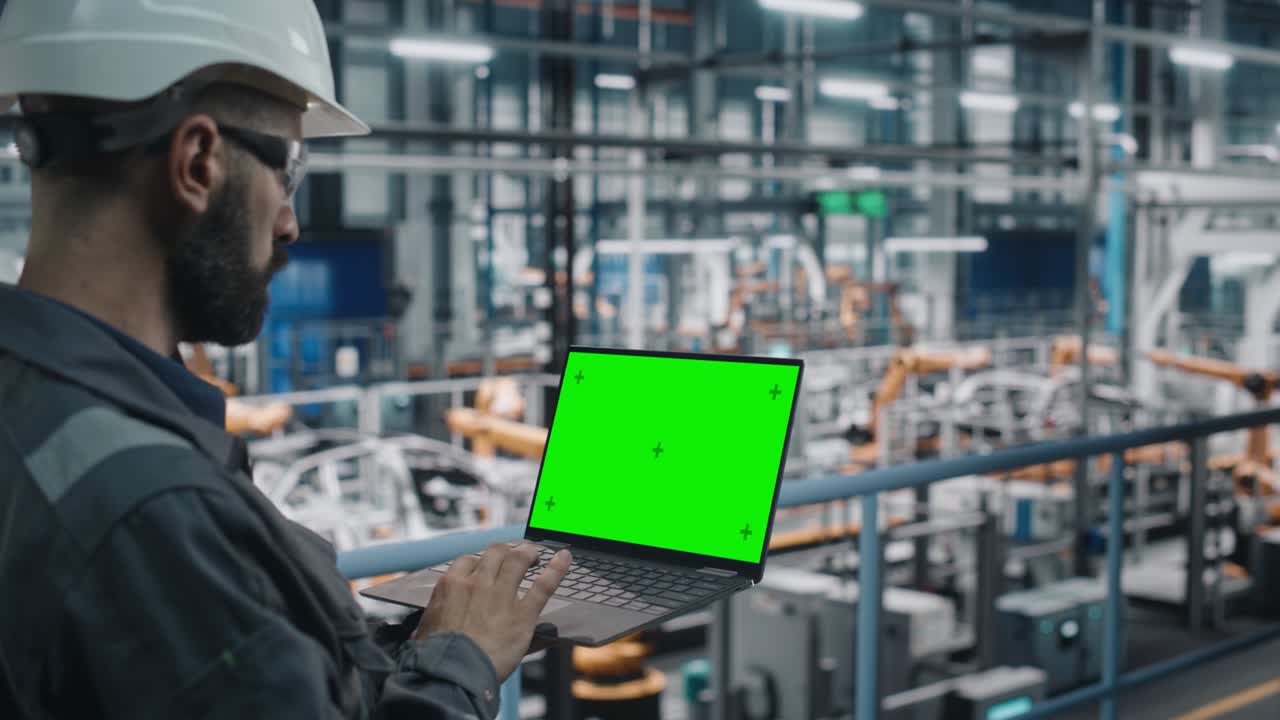작업 유니폼을 입은 자동차 공장 엔지니어는 녹색 화면 모 디스플레이를 가진 노트북 컴퓨터를 사용하여 차량 생산에 전념하는 자동차 산업 제조 시설에서 소프트웨어를 사용합니다.