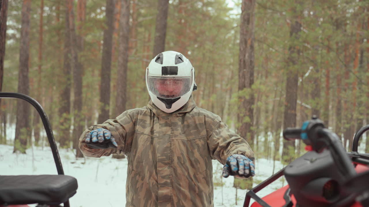 Un uomo prepara un quad rosso in una foresta di pini innevata, uno scout fa segnali come un ranger forestale, ispeziona motore e attrezzatura, sale sul veicolo in mimetica e casco, avvia l'acceleratore e guida.
