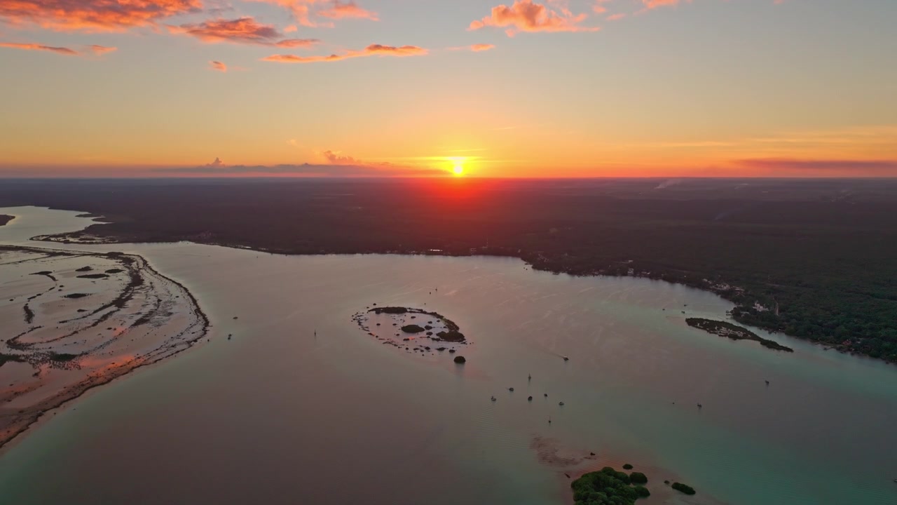 Ilmakuva hehkuvasta auringonlaskusta hiekkasärkän yllä Bacalarin laguunissa, Meksikossa, pastellivalon heijastuessa tyynestä ja peilityynestä vedestä.