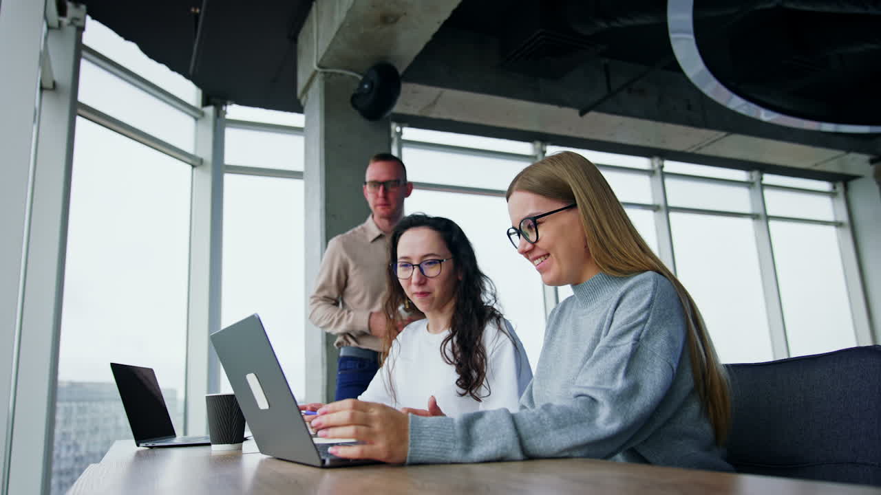 Happy positive people laugh looking at laptop. Male colleague comes up standing behind the ladies. Low angle view.