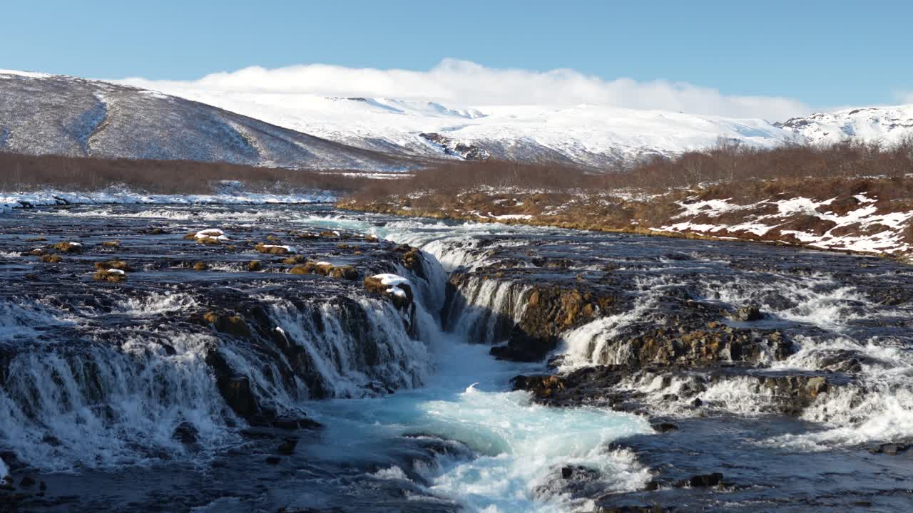 Aerial view of Brúarfoss waterfall in Iceland, where icy blue glacial waters cascade over dark volcanic rocks, surrounded by snow-dusted terrain and distant winter hills.