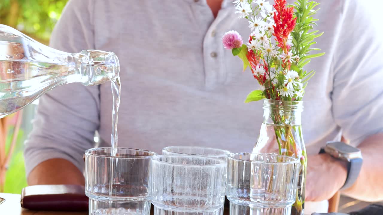 A man in a light shirt pours water from a glass carafe into a tumbler while holding a menu at a sunlit outdoor café table with floral centerpiece
