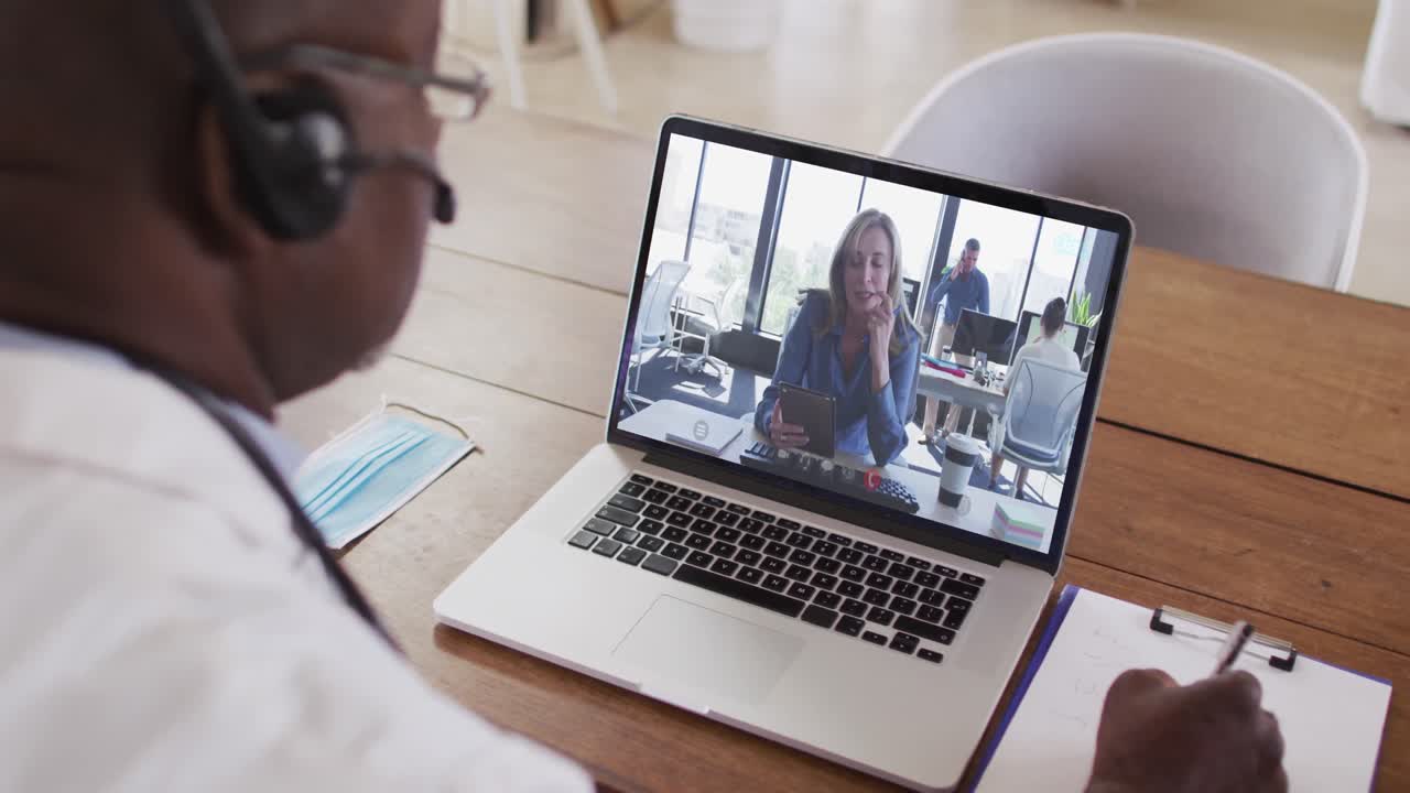 Afroamerikansk mandlig læge med headset på, der tager noter under et videoopkald på sin bærbare computer