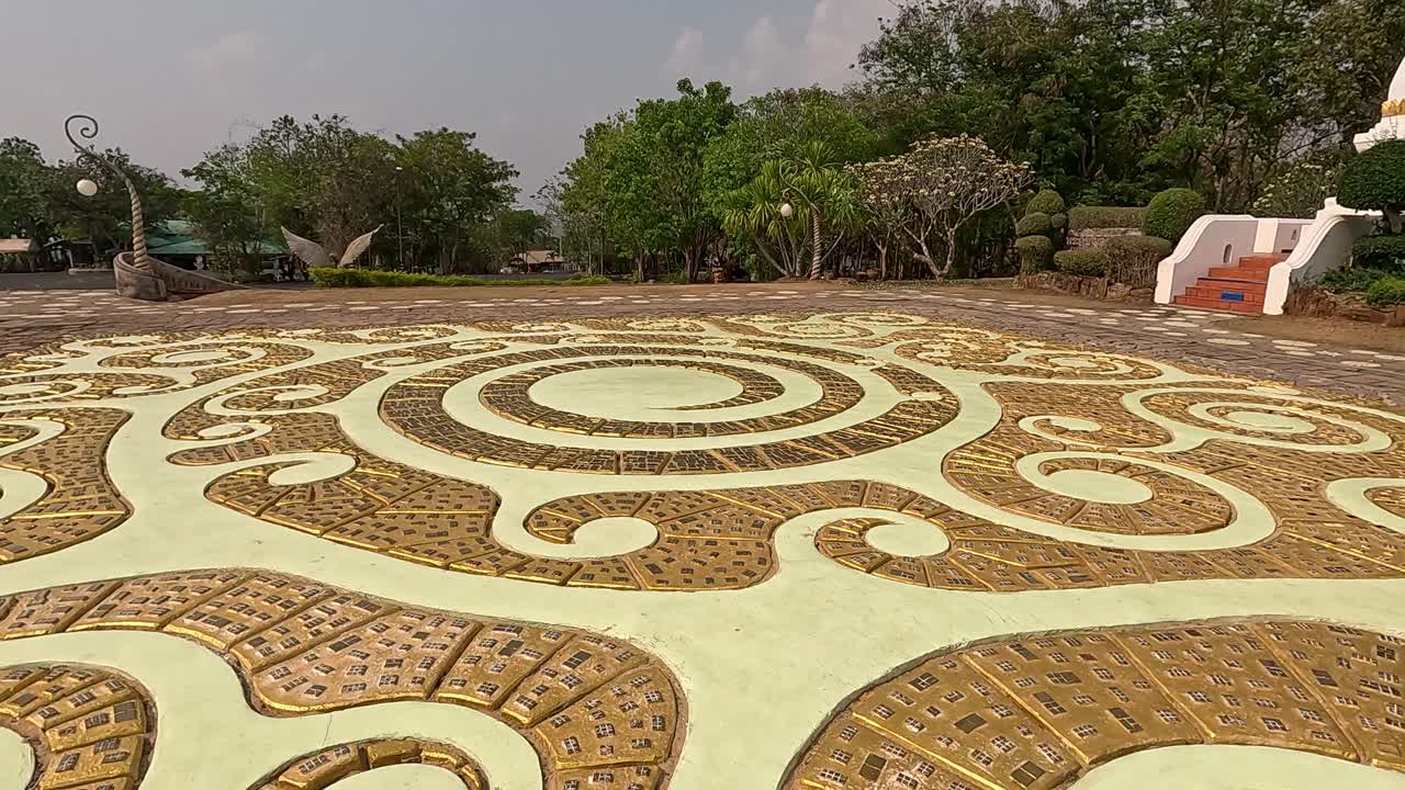 Low-angle camera pans across ornate circular mosaic tiles in an outdoor Buddhist temple courtyard, with greenery and a white stupa visible under soft daylight