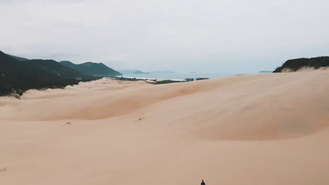 Establishing-shot-of-sandboarder-walking-on-sand-dunes-at-Garopaba-Beach,-Santa-Catarina,-Brasil