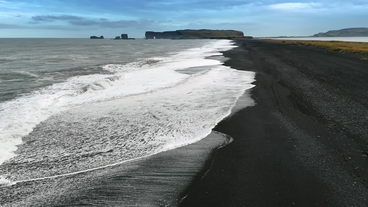 Sea waves splashing on the beach with black sand. Silhouettes of steep rocks at backdrop. Iceland wilderness.