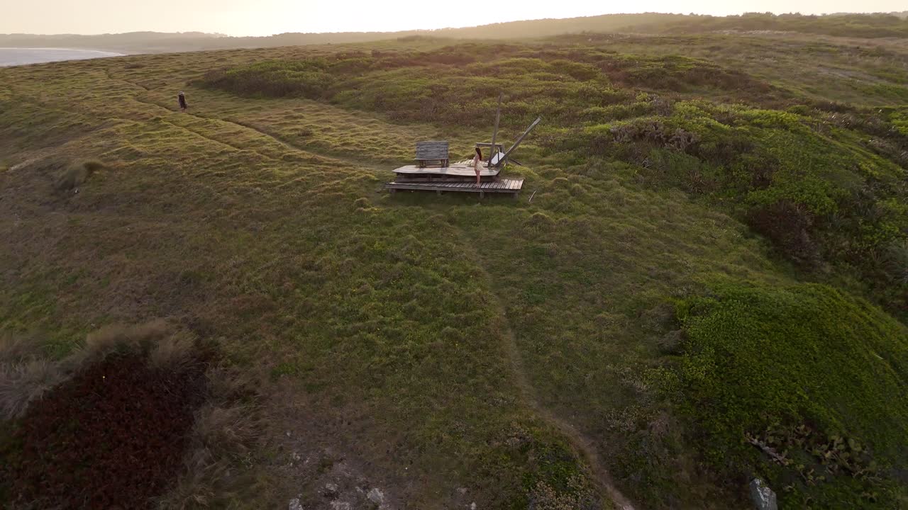Cerro Chato no Parque Santa Teresa, Uruguai, ao pôr do sol, com mulher e homem reunidos em plataforma de madeira com vista para o Oceano Atlântico em luz dourada quente. Tomada aérea orbital
